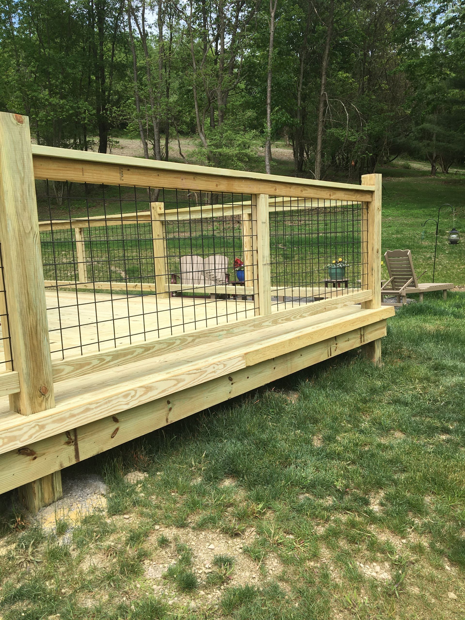 A wooden deck featuring a railing with dark, vertical metal balusters, set in a grassy outdoor area near trees.