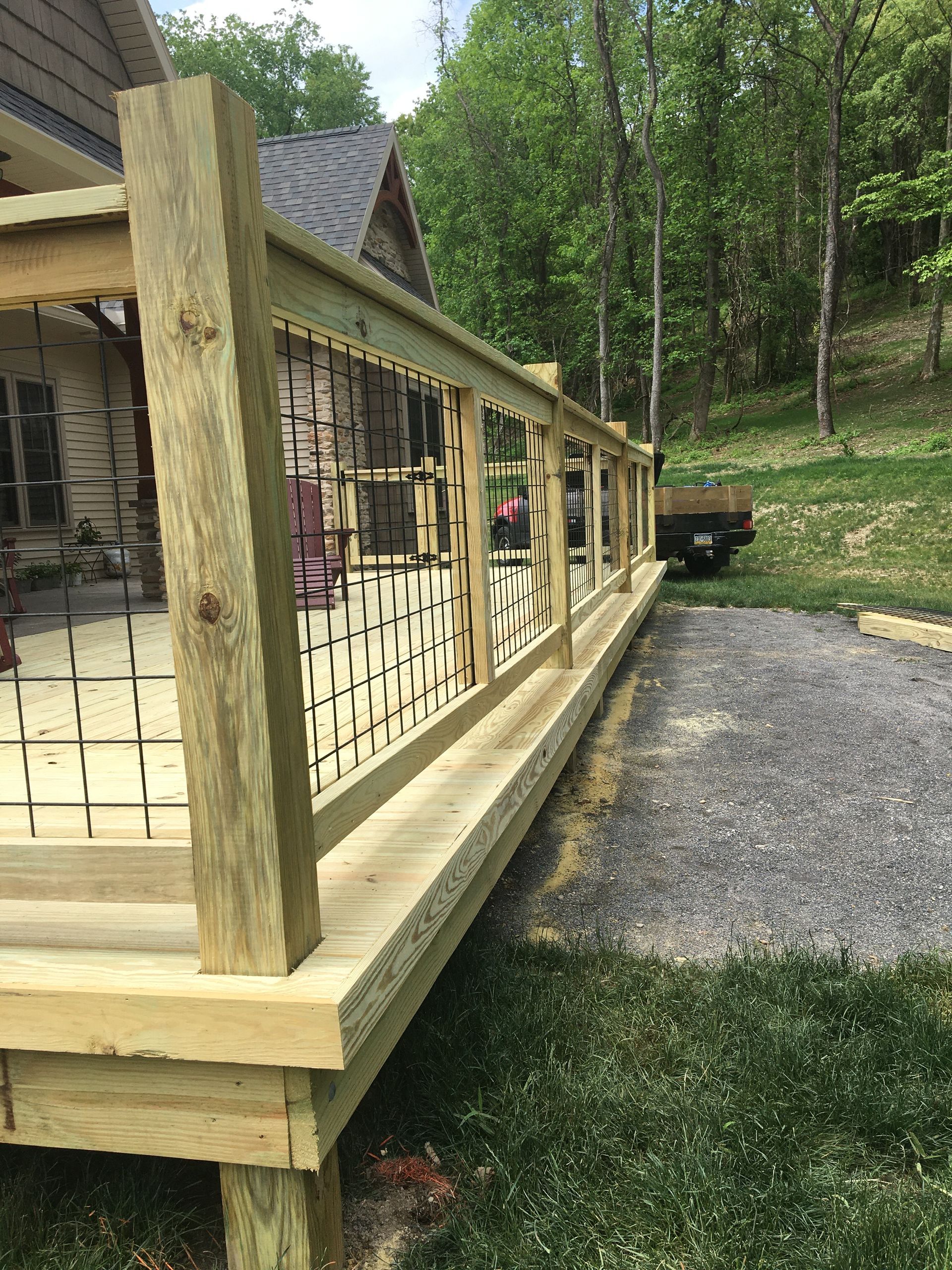 A wooden deck with a wire grid railing, built outdoors near a house and a gravel driveway on a sunny day.