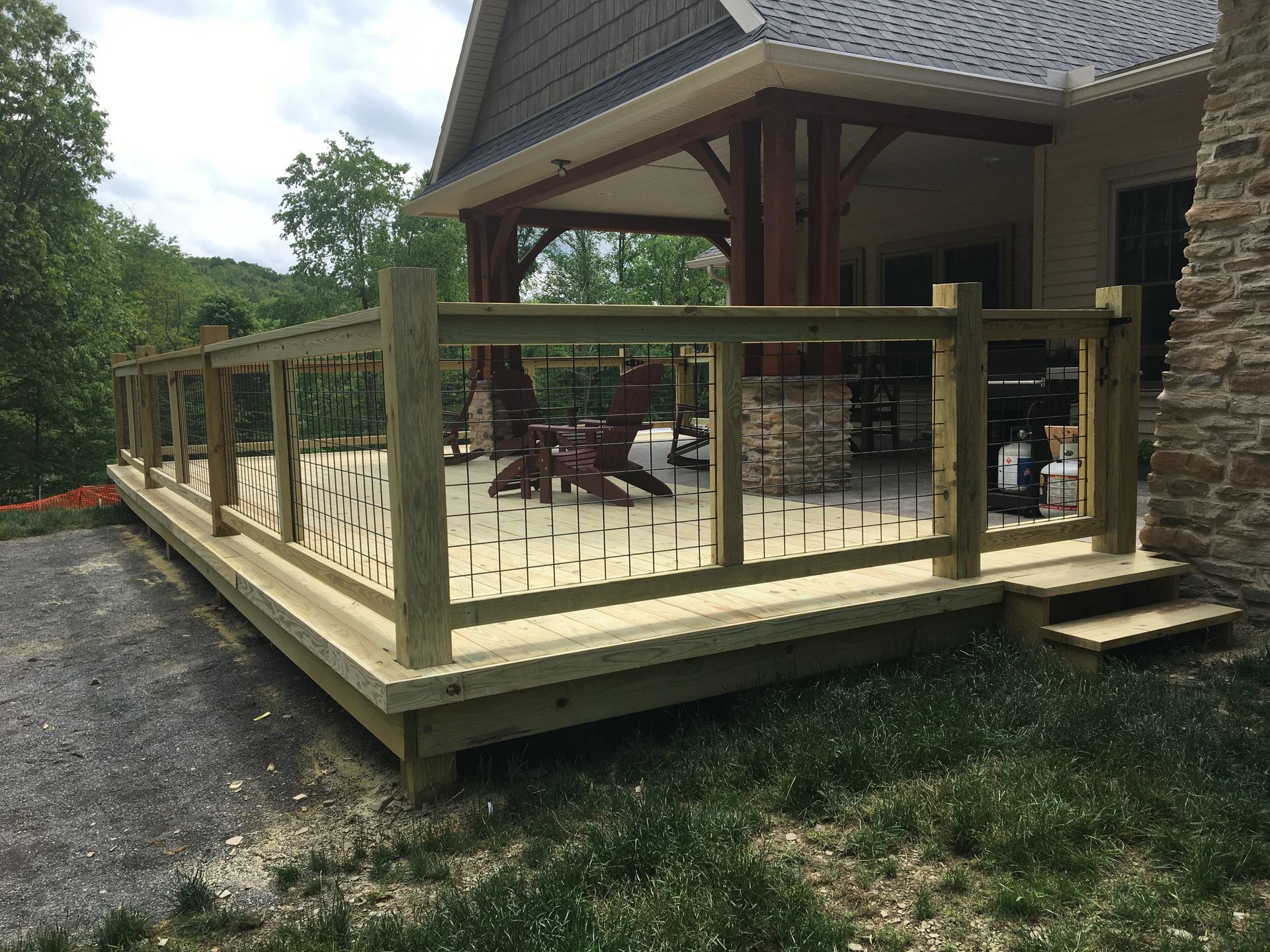 A wooden deck with metal railing, chairs, and a covered porch area attached to a house with stone siding.