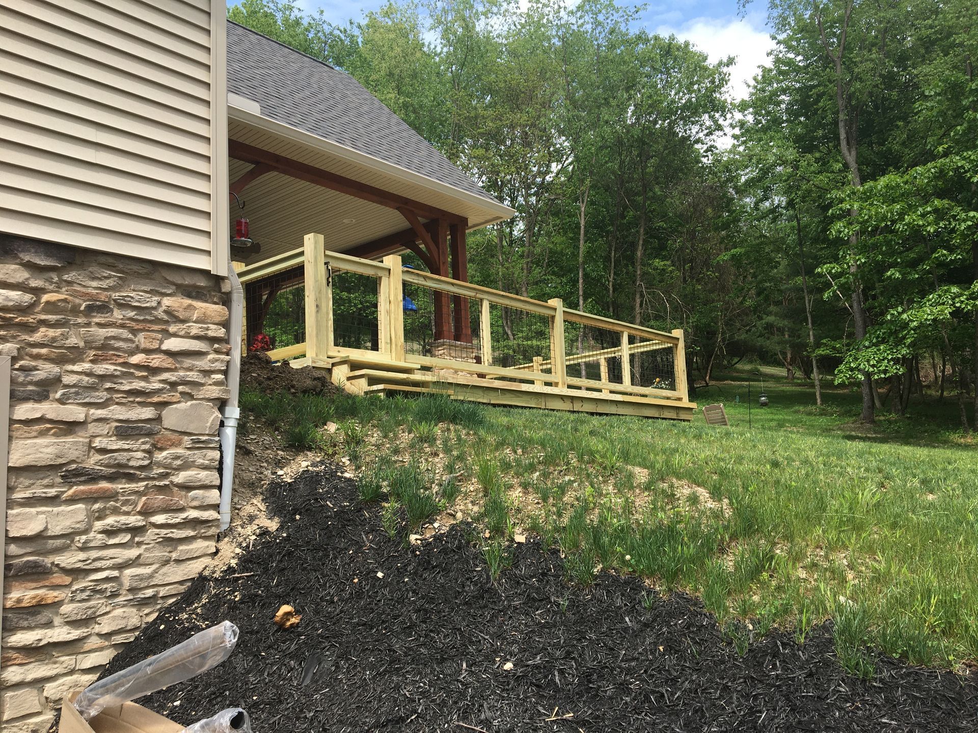 A wooden wheelchair ramp with railings extending from a stone and siding house into a grassy, wooded backyard.
