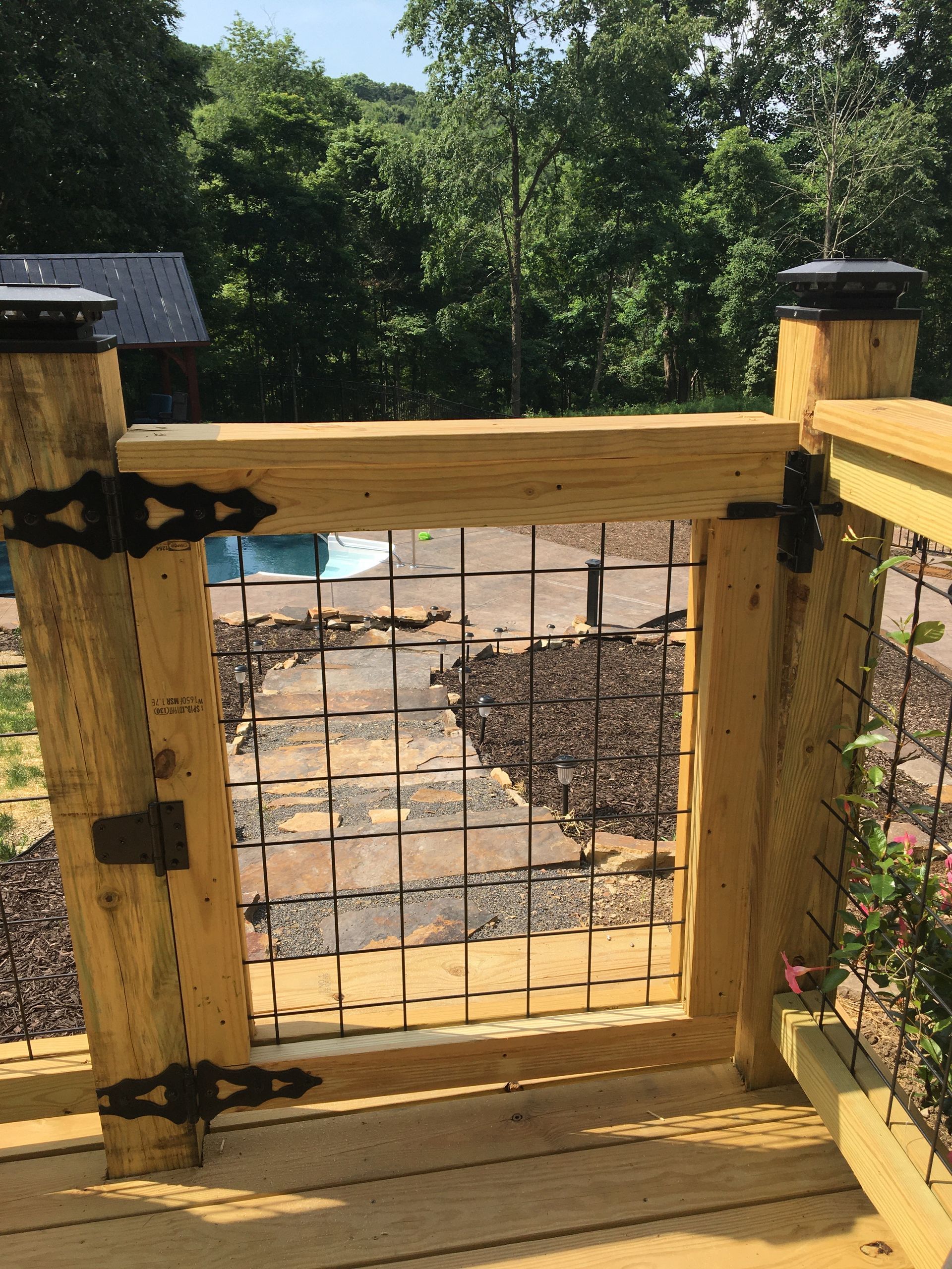 A wooden gate with a wire mesh panel installed on a deck, looking out toward a backyard path and trees.