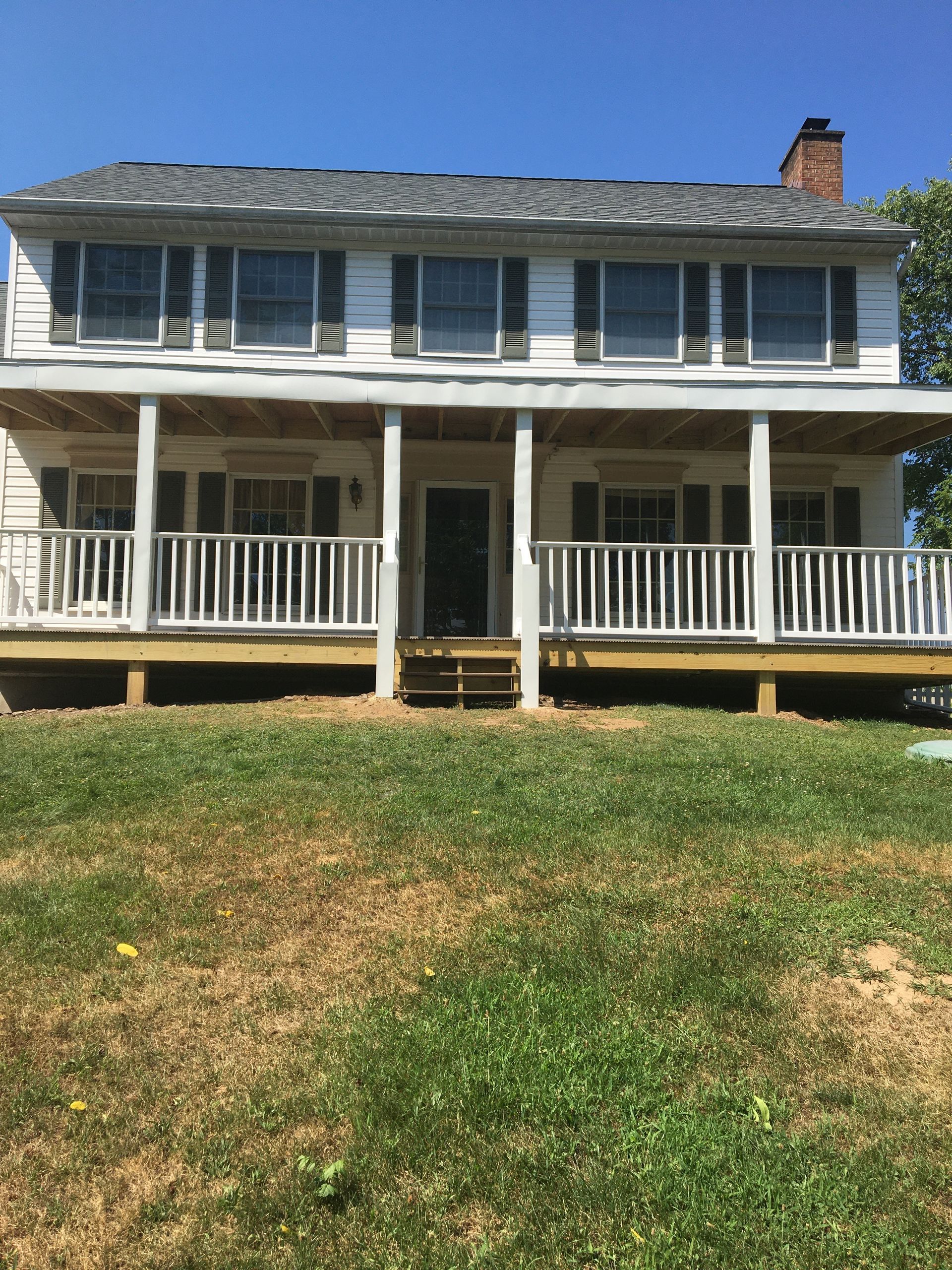 A two-story white house with a large front porch, black shutters, and a chimney, situated behind a grassy yard.
