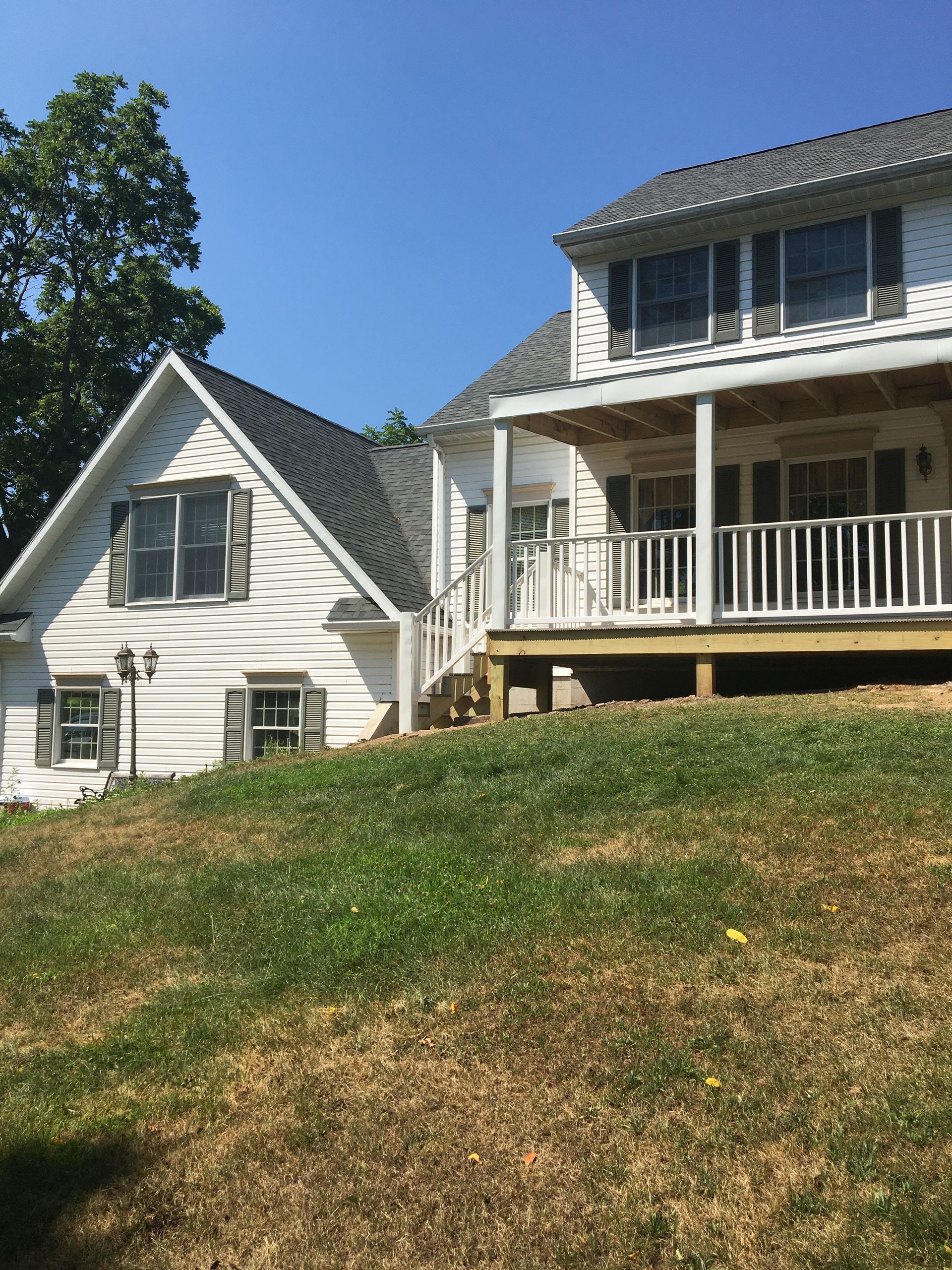 A two-story white house with dark shutters and a wooden deck sits on a grassy hill under a clear blue sky.