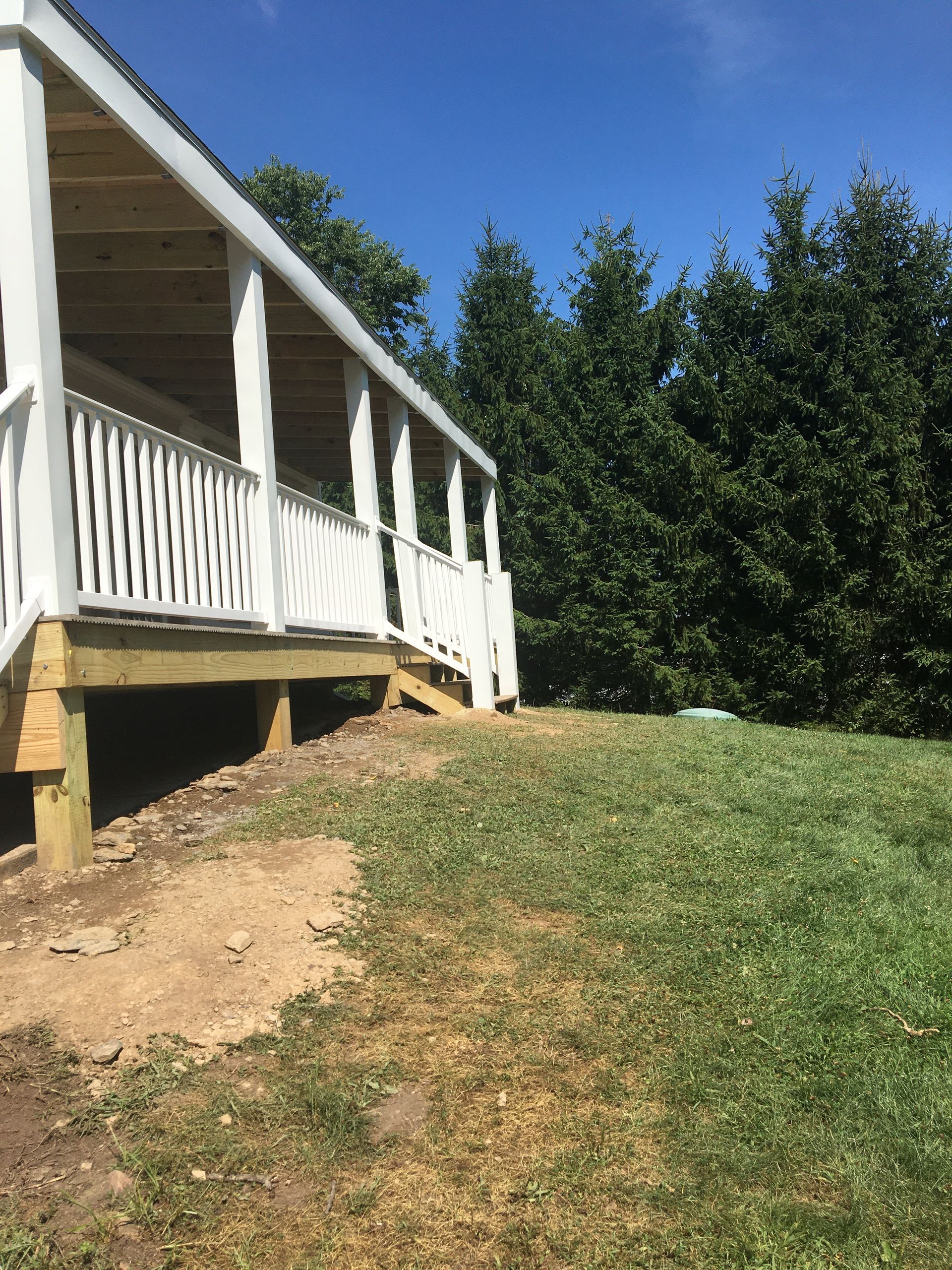 A wooden porch with white railings attached to a house, set above a grassy, slightly sloping yard with tall evergreen trees.