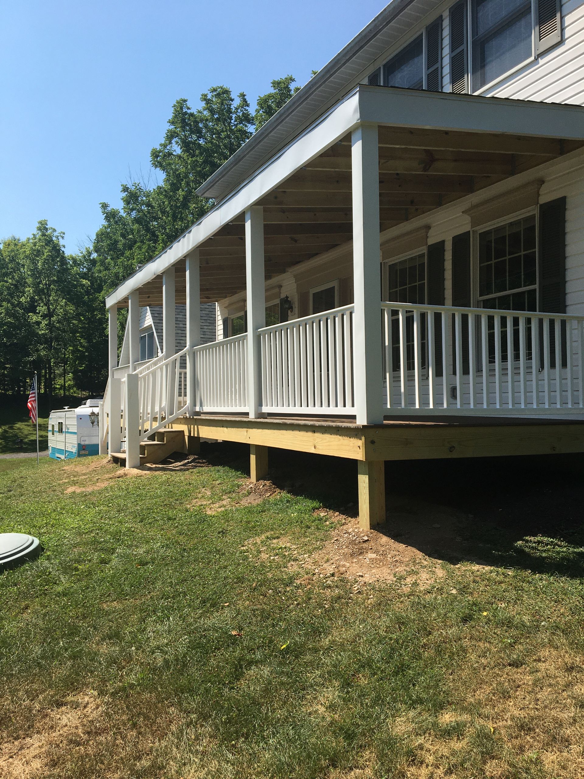 A white house with a covered wooden porch, featuring white railings and support posts, set on a grassy lawn.