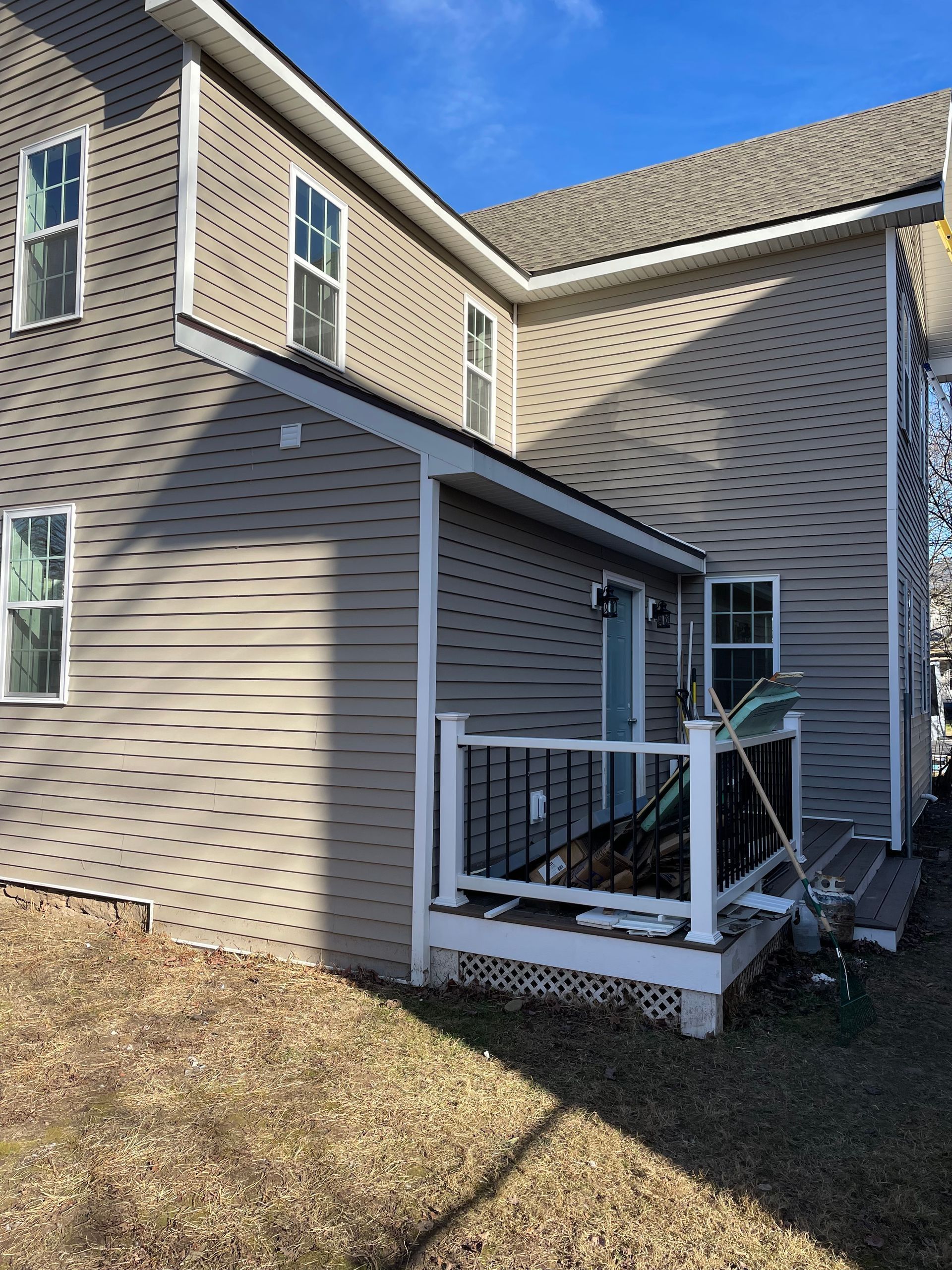 A two-story tan house with a small side deck, white railings, and a blue door under a clear blue sky.
