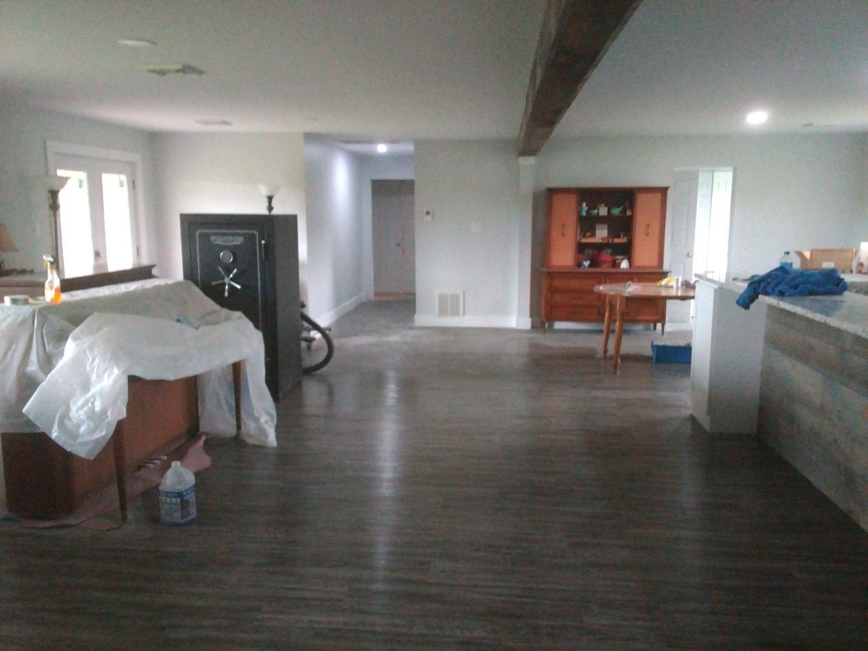 An open-plan room with wood-look flooring, a large black gun safe, a covered piece of furniture, and a distant hutch.