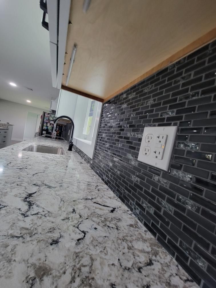 A kitchen backsplash of dark grey subway tiles above a white marble countertop with an electrical outlet.