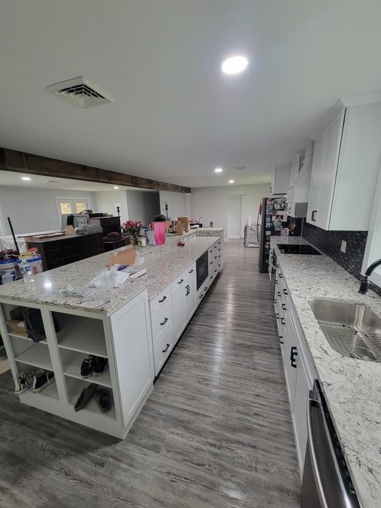 A wide-angle view of a modern kitchen with a long granite-topped island, white cabinets, and wood-look laminate flooring.