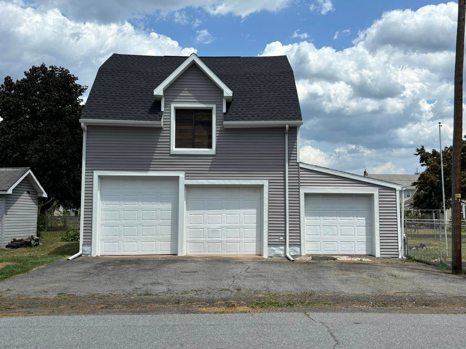 A gray two-story detached garage with a dark roof and three white garage doors set on a gravel lot under a cloudy sky.