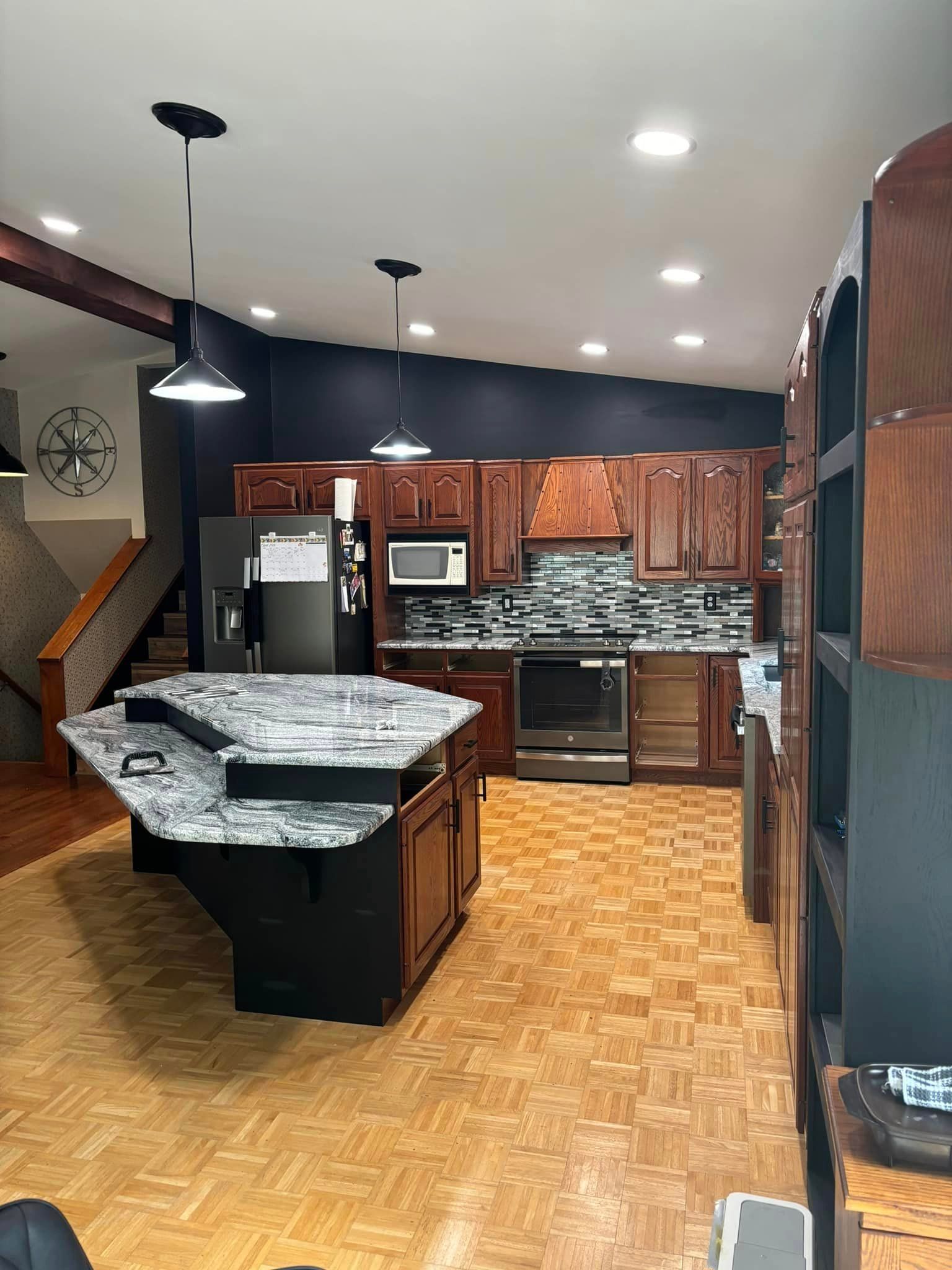 A modern kitchen with black cabinets, a granite-topped island, a tiled backsplash, and parquet flooring.