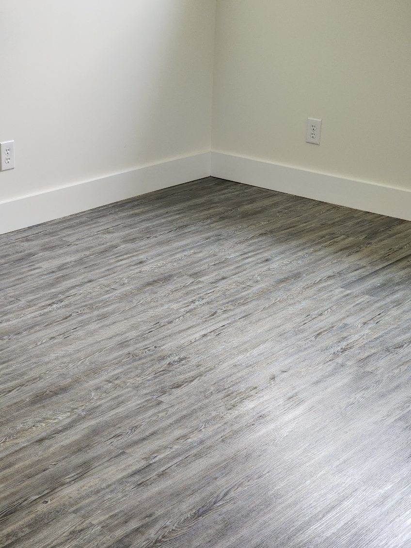 The corner of a room featuring grey wood-look plank flooring, white baseboards, and plain, light-colored walls.