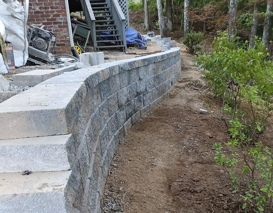 Stone retaining wall curving along a dirt path, with stairs on the left and trees in the background.