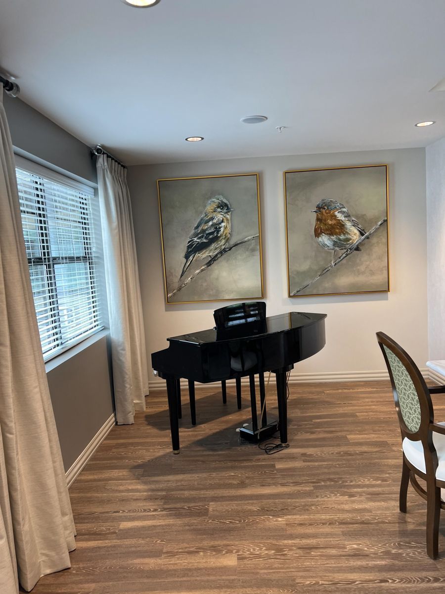 A black baby grand piano sits beneath bird paintings. The room has curtains, a window, and a dining chair.
