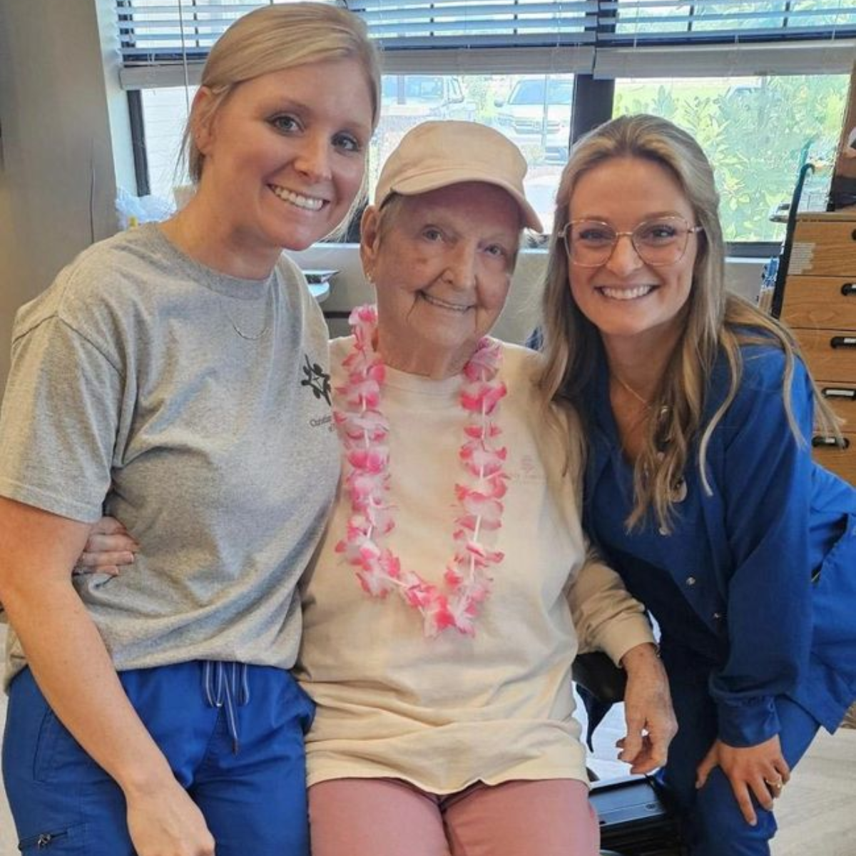 Three smiling women pose indoors; a senior woman wears a floral lei, flanked by two younger women.