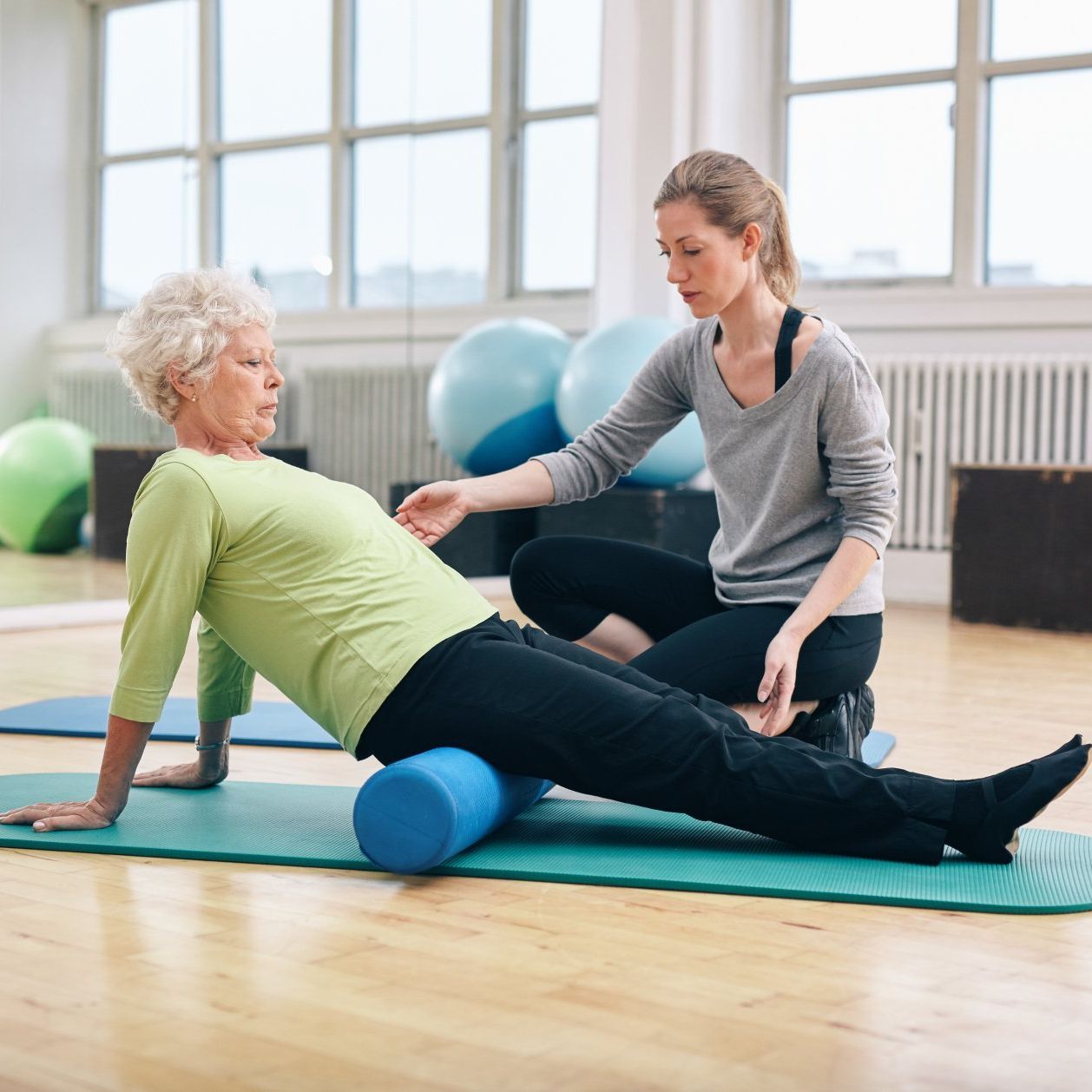An older woman exercises with trainer using a foam roller in a gym.