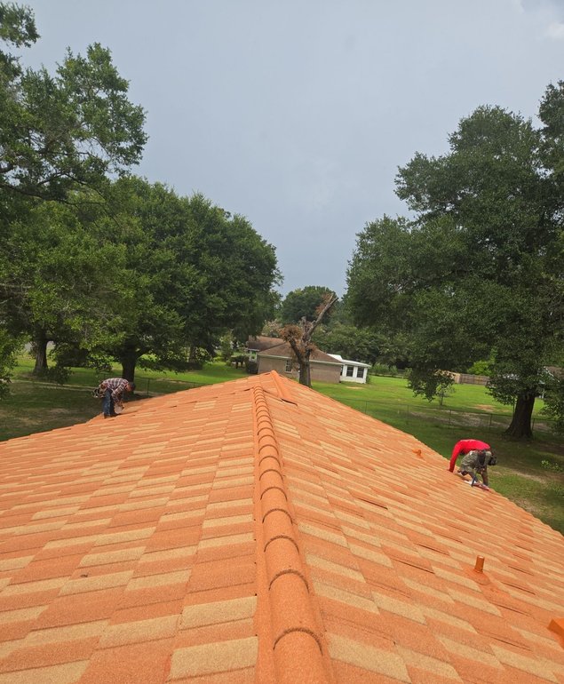 Roofers installing new orange and tan shingles on a house roof. Green trees and overcast sky in the background.