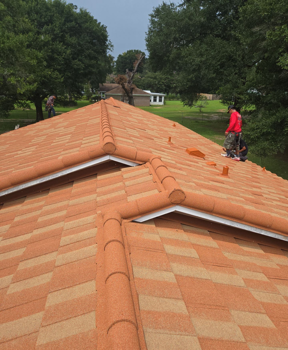 Roofers on a residential roof with brown and tan tiles, surrounded by trees.