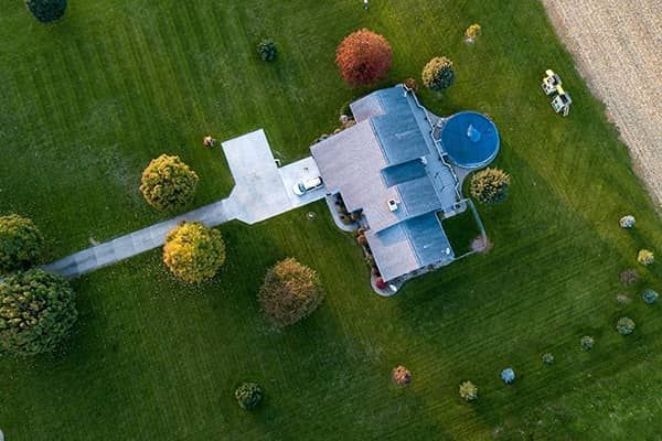 Aerial view of a house with a driveway, surrounded by green grass and trees, and a blue circular pool.
