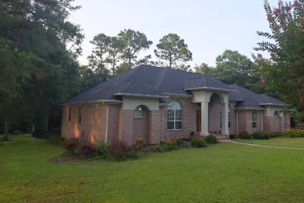 Brick house with dark roof and white columns, surrounded by trees and green lawn.