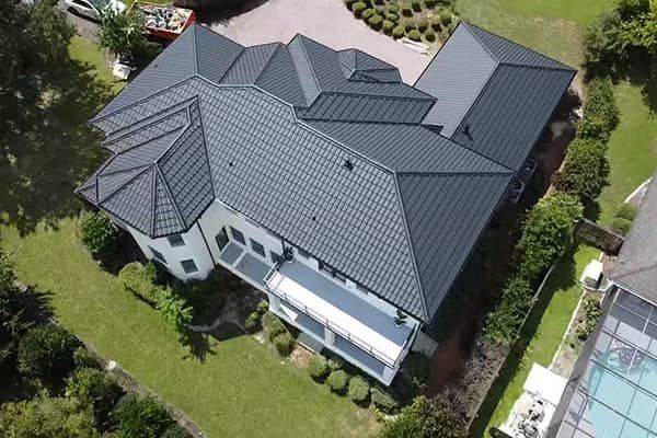 Aerial view of a large, modern home with a dark gray metal tile roof and a surrounding green lawn.
