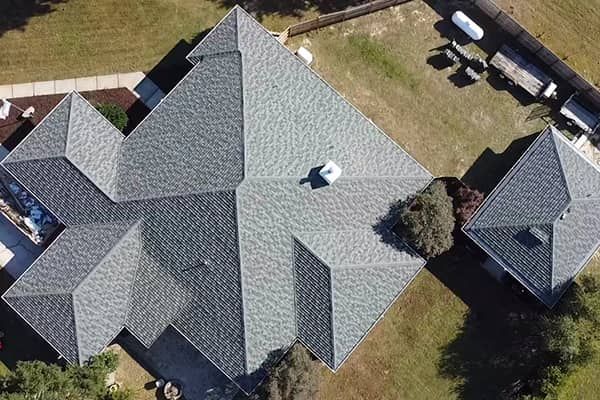 Overhead view of a house with a complex, gray shingle roof, surrounded by lawn and some trees.
