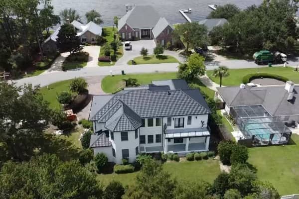 Aerial view of a large white house with a dark gray roof, surrounded by trees and other houses.