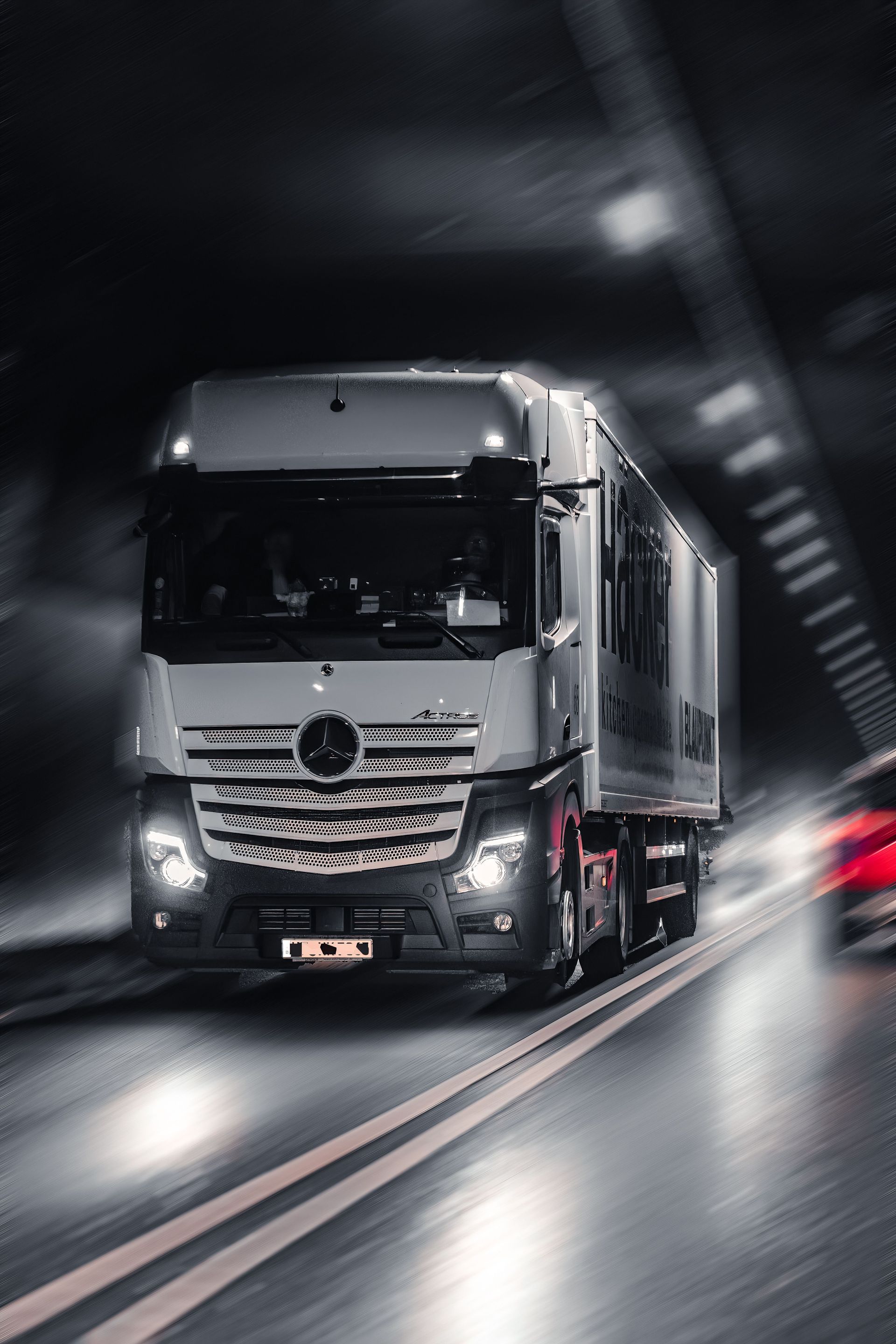 Een witte vrachtwagen raast met hoge snelheid door een schemerige tunnel, koplampen aan, bewegingsonscherpte en rode achterlichten in beeld.