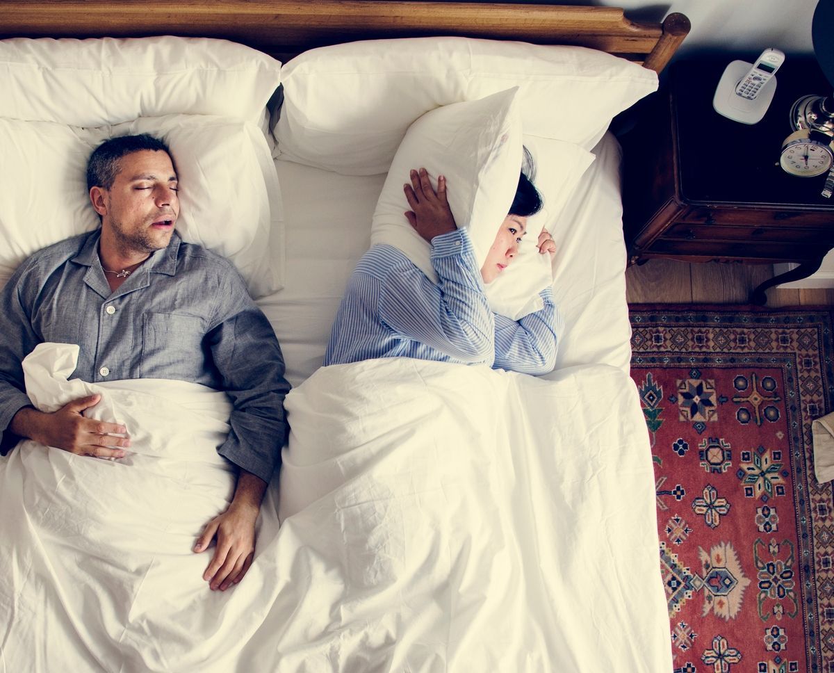 A man and woman are in bed with the man snoring loudly and the woman covering her head with a pillow
