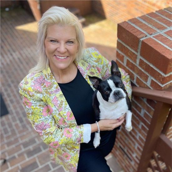 A blonde-haired smiiling woman is standing holding a Boston Terrier dog with a floral blazer on.