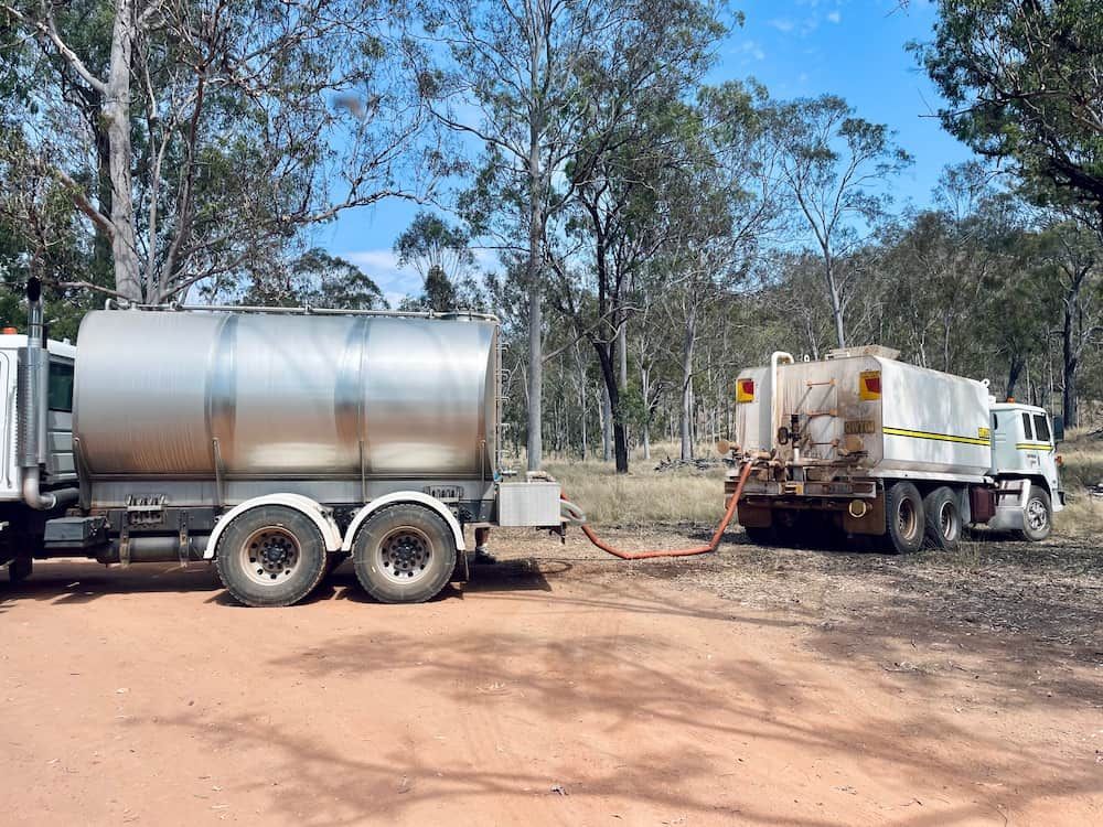 Two Tanker Trucks Connected by a Hose on a Dirt Road, Trees — LS Plant Hire in Calliope, QLD