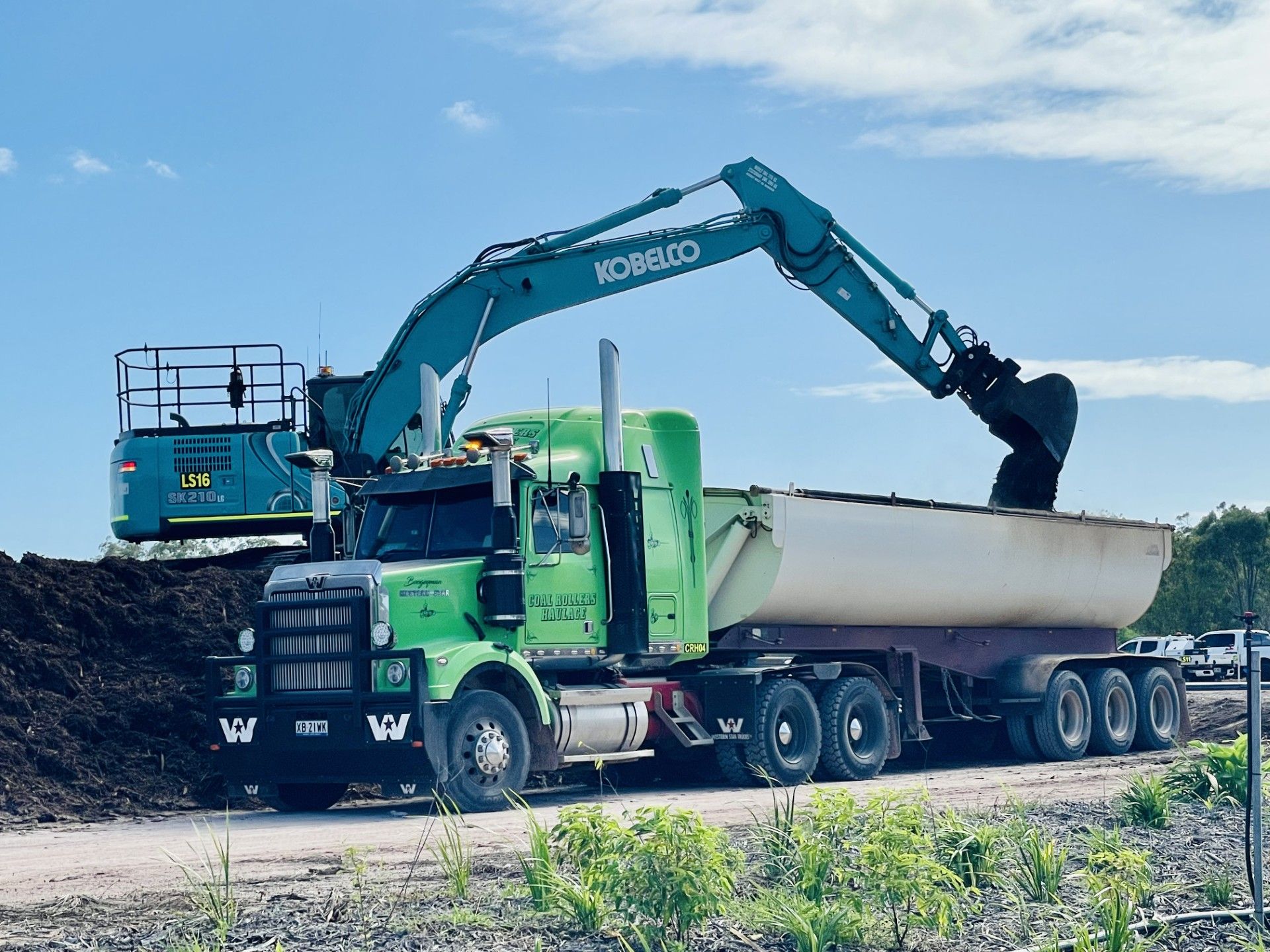 A green semi-truck being loaded with soil by a teal Kobelco excavator on a construction site under a blue sky. — LS Plant Hire in Calliope, QLD