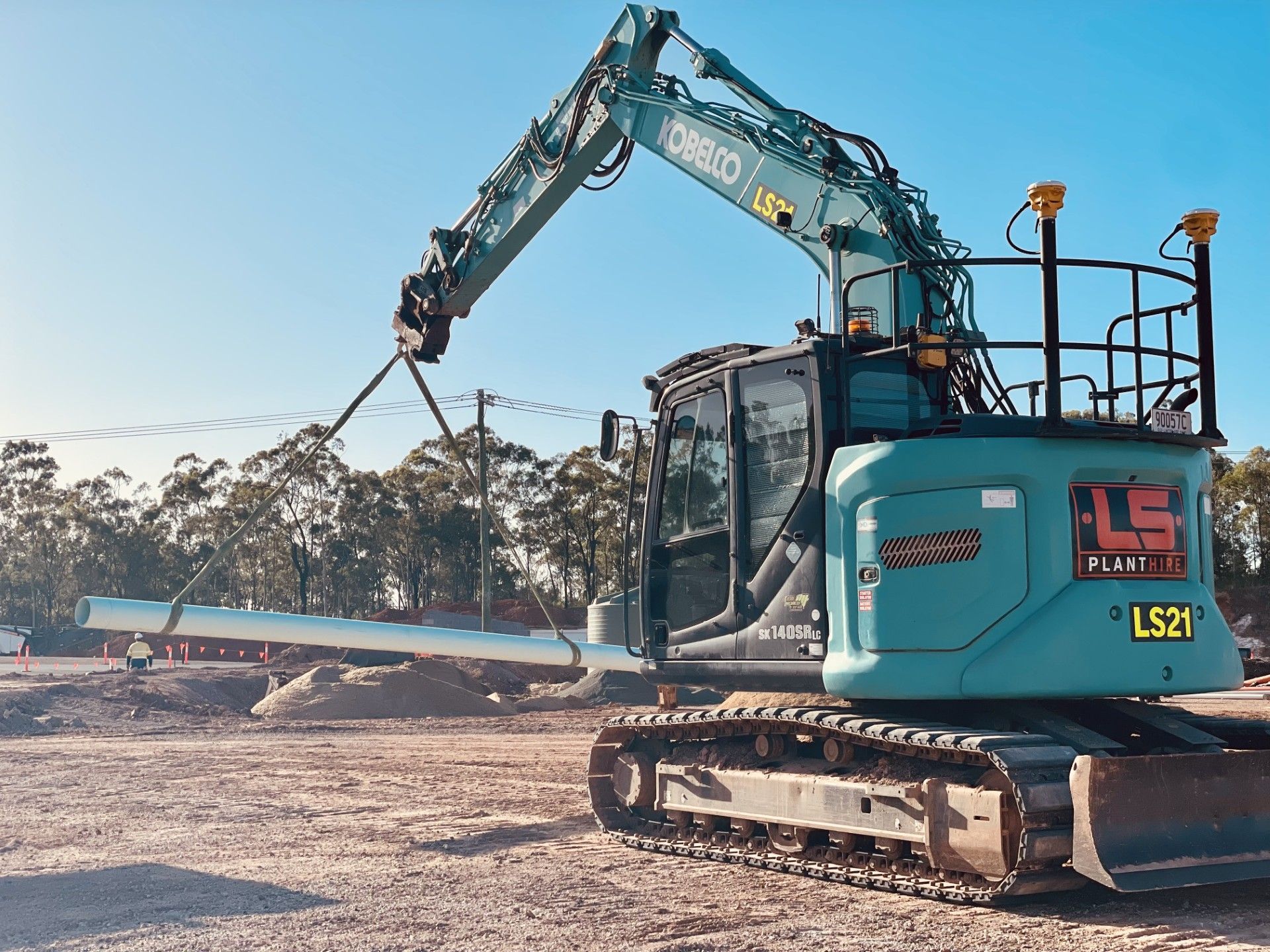 A teal Kobelco excavator lifts a long pipe on a dirt construction site under a clear blue sky. — LS Plant Hire in Calliope, QLD