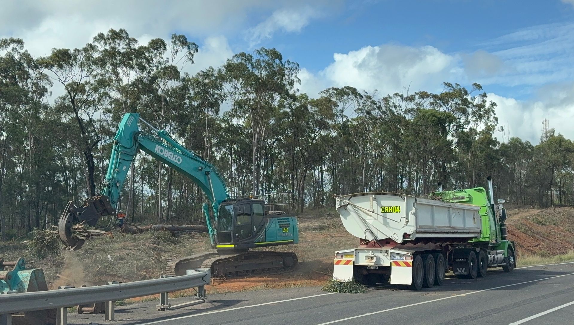 A turquoise excavator uses a grapple attachment to load debris into a dump truck parked beside a forested roadside. — LS Plant Hire in Calliope, QLD