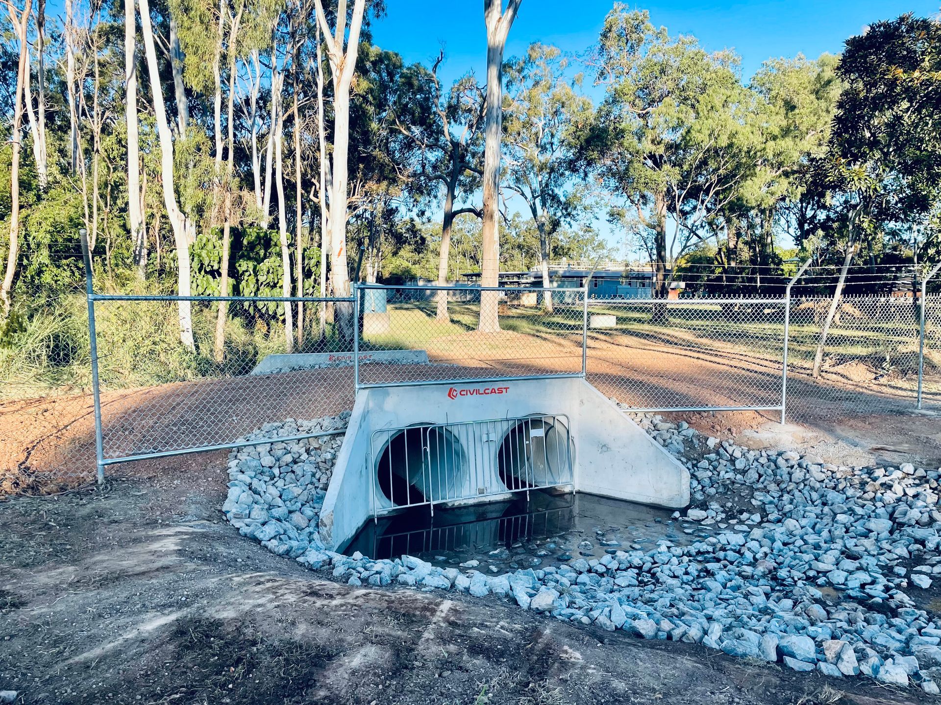 A double concrete storm drain pipe with a metal grate set in a dirt clearing surrounded by trees and a wire fence. — LS Plant Hire in Calliope, QLD