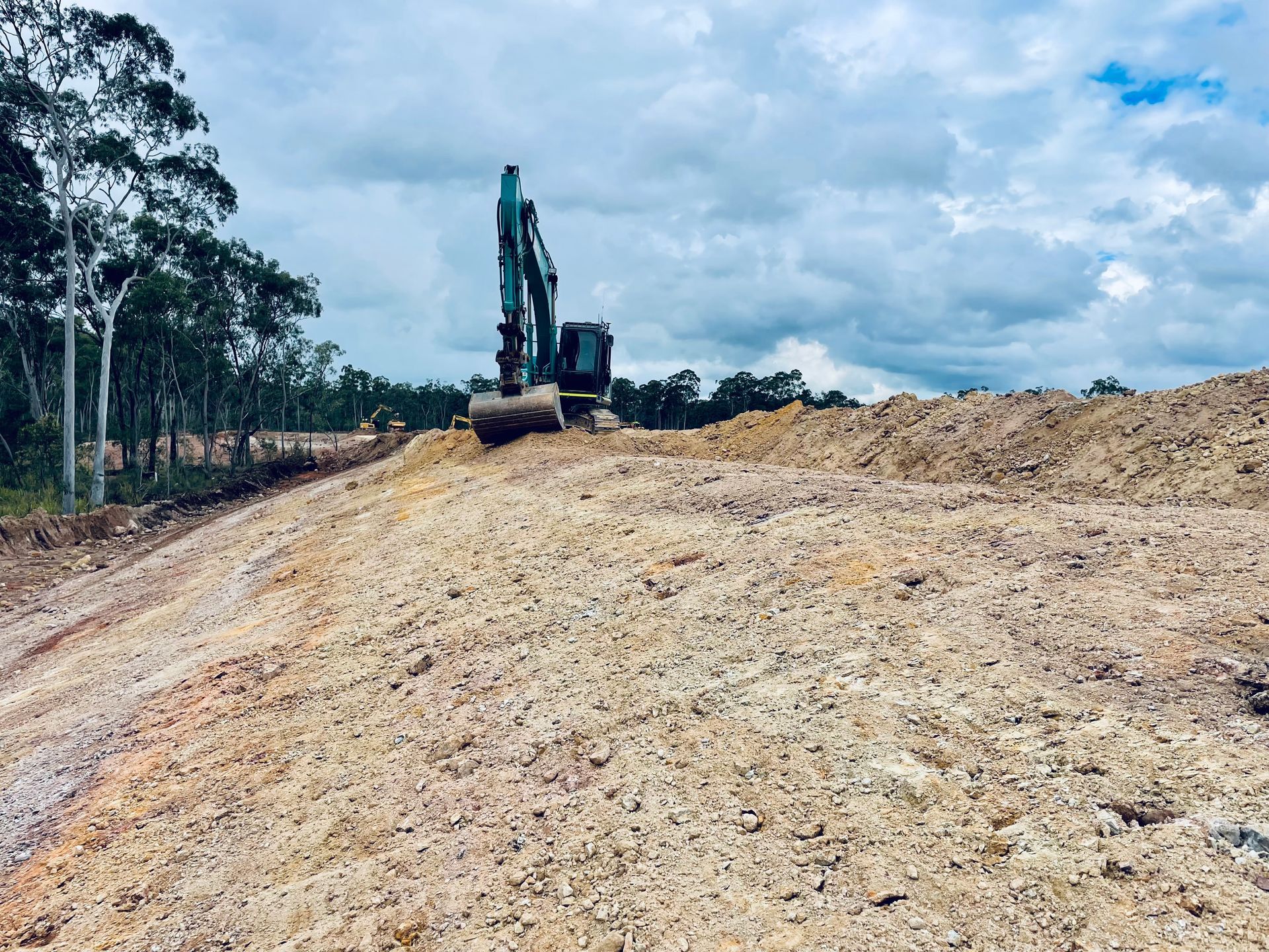 An excavator sits atop a large mound of loose, light-colored dirt under a cloudy sky near a line of trees. — LS Plant Hire in Calliope, QLD