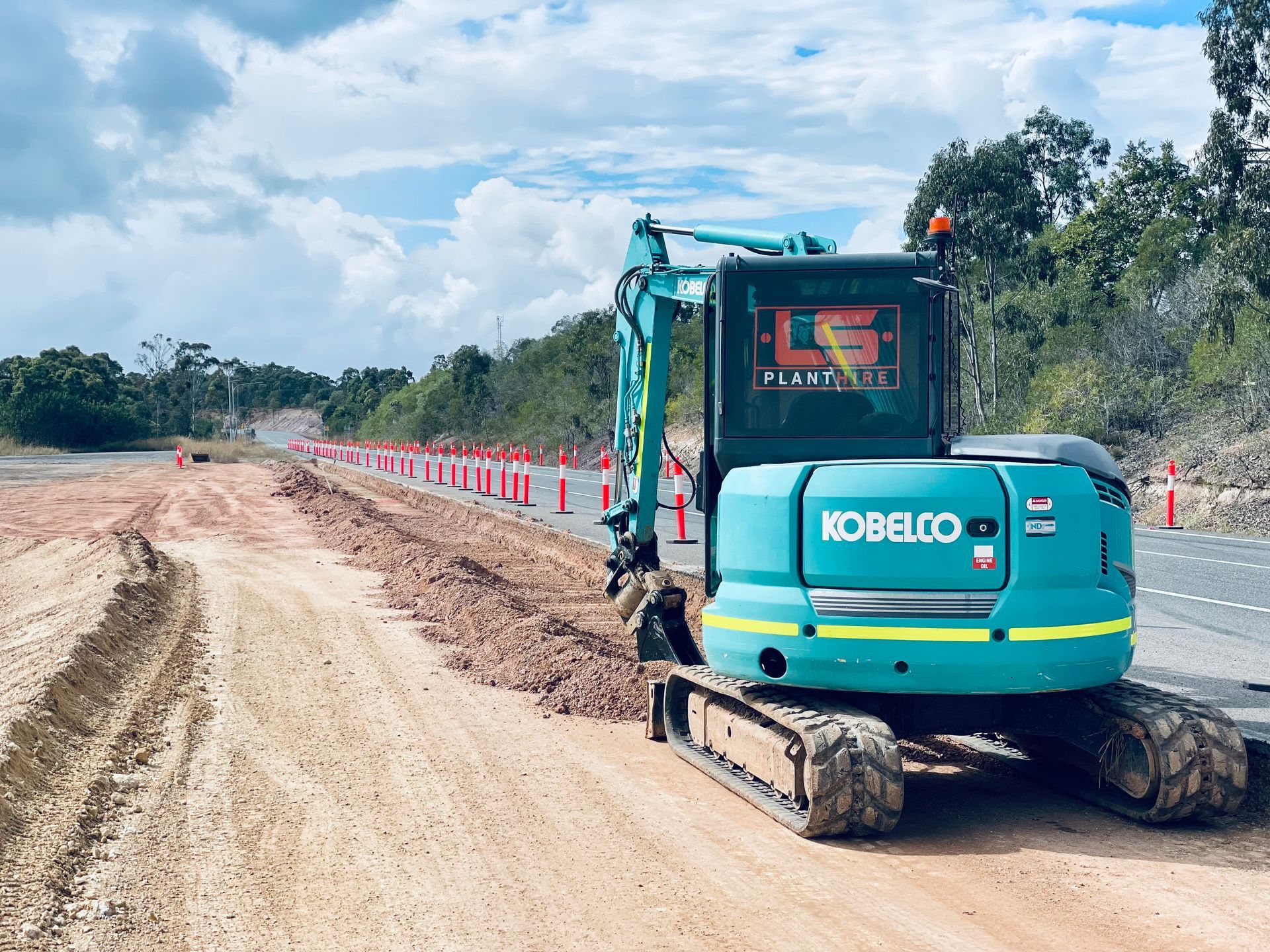 A teal Kobelco excavator working on a roadside dirt embankment next to a paved road lined with orange traffic cones. — LS Plant Hire in Calliope, QLD