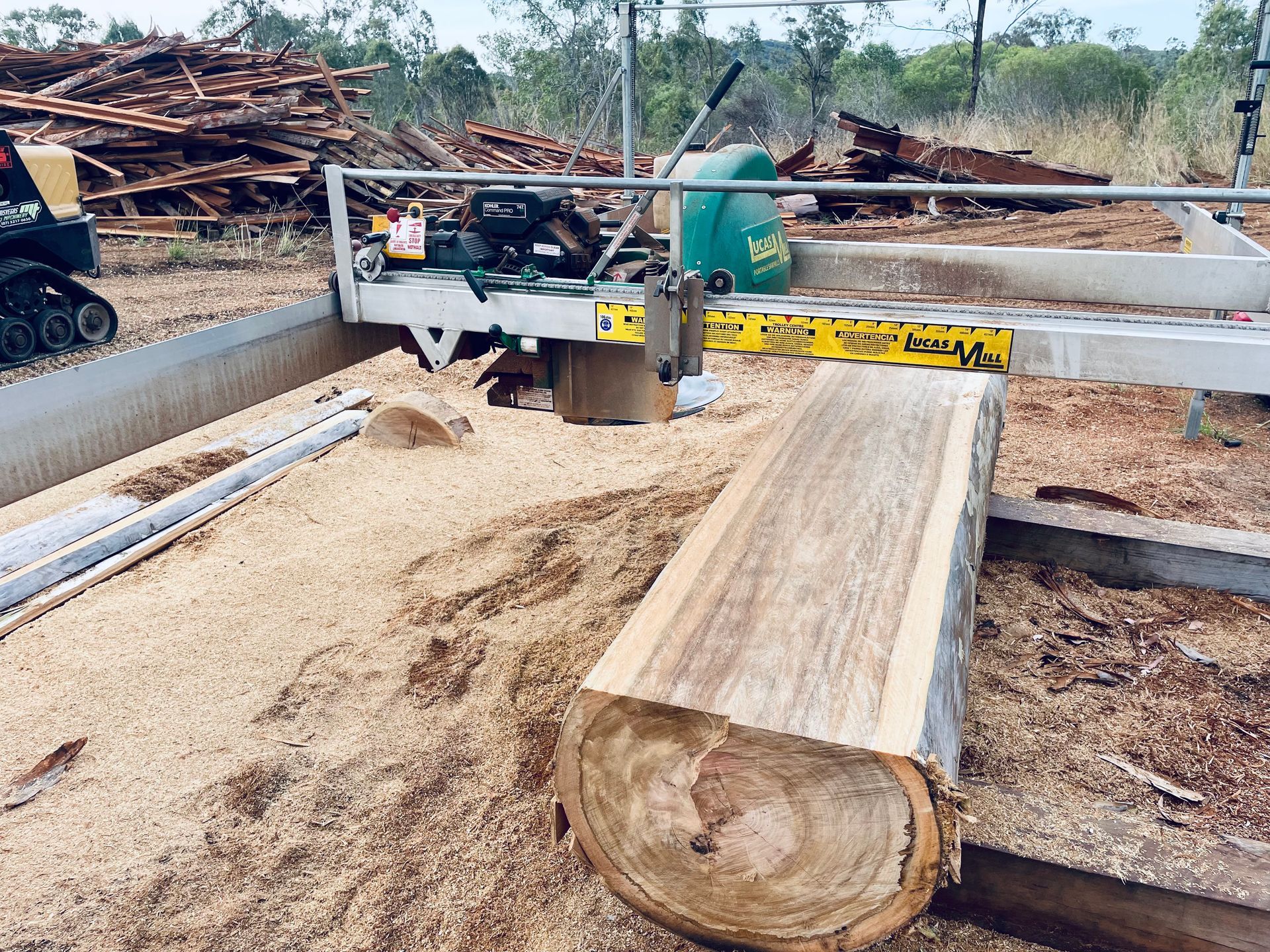A portable sawmill processes a large wooden log in a outdoor, sawdust-covered field filled with stacked timber. — LS Plant Hire in Calliope, QLD