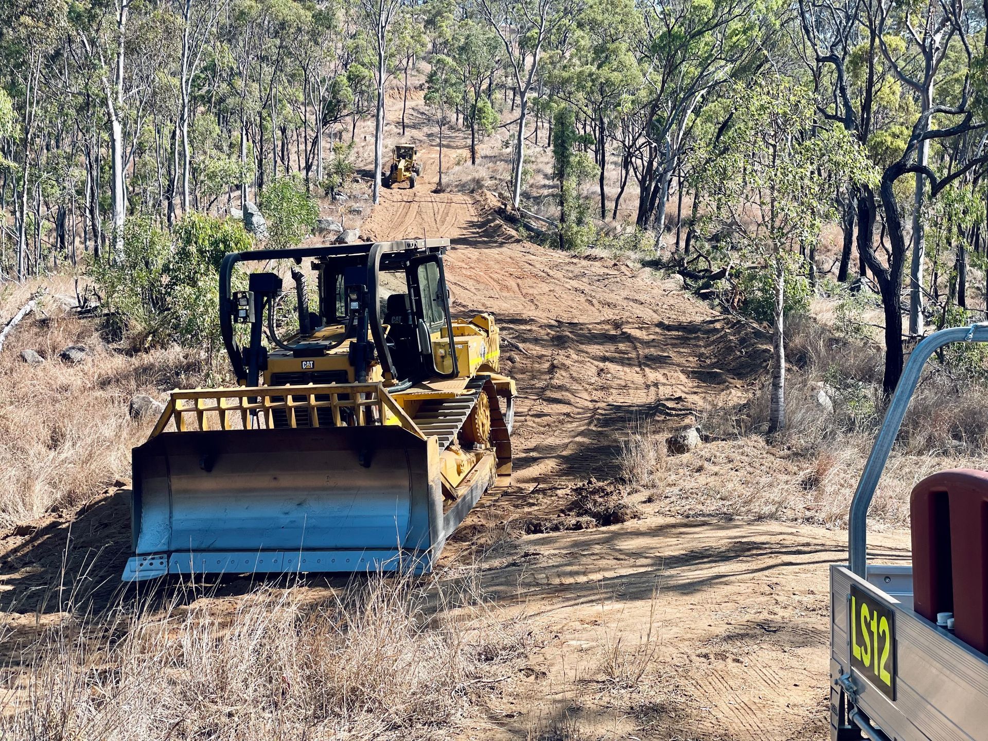 A yellow bulldozer drives up a dirt path in a wooded area, with another piece of machinery visible further up the hill. — LS Plant Hire in Calliope, QLD