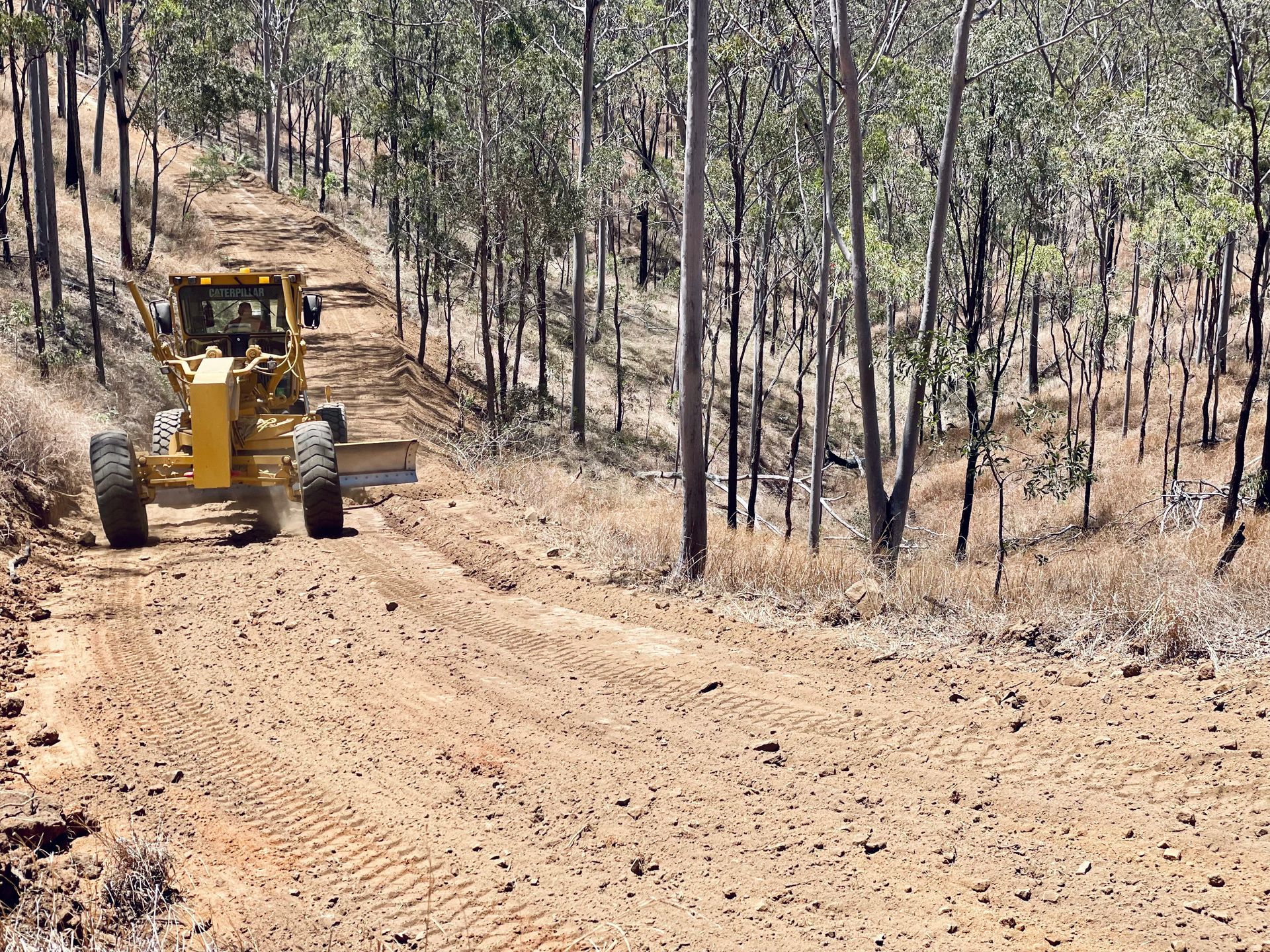 A yellow motor grader smoothing a dirt road through a sparsely forested, hilly landscape. — LS Plant Hire in Calliope, QLD