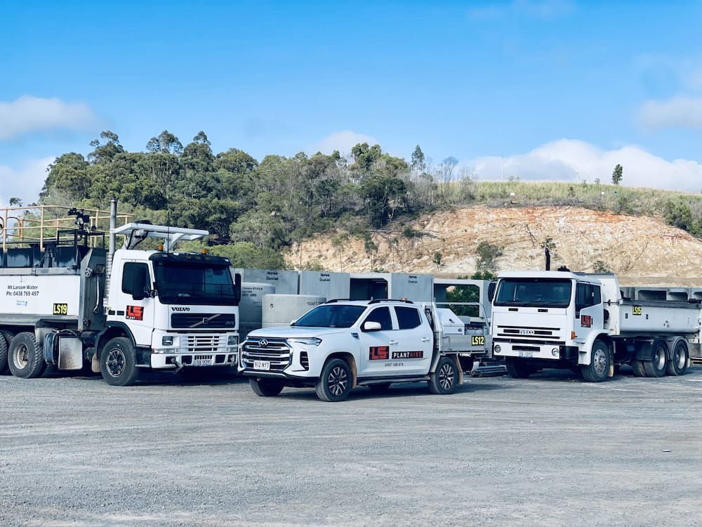 Three White Trucks and a White Pickup Truck Parked on Gravel in Front of a Hillside — LS Plant Hire in Calliope, QLD