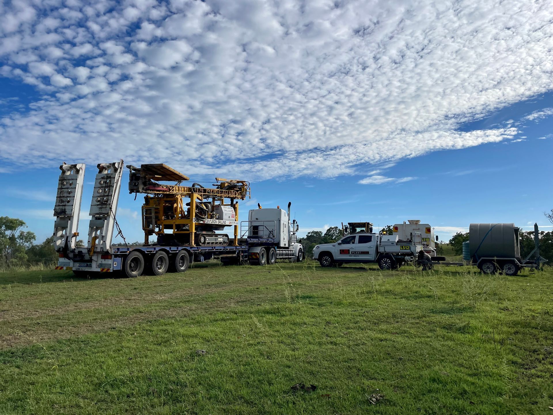 Heavy machinery loaded on a flatbed trailer in a grassy field with a support truck and equipment trailer under blue skies. — LS Plant Hire in Calliope, QLD