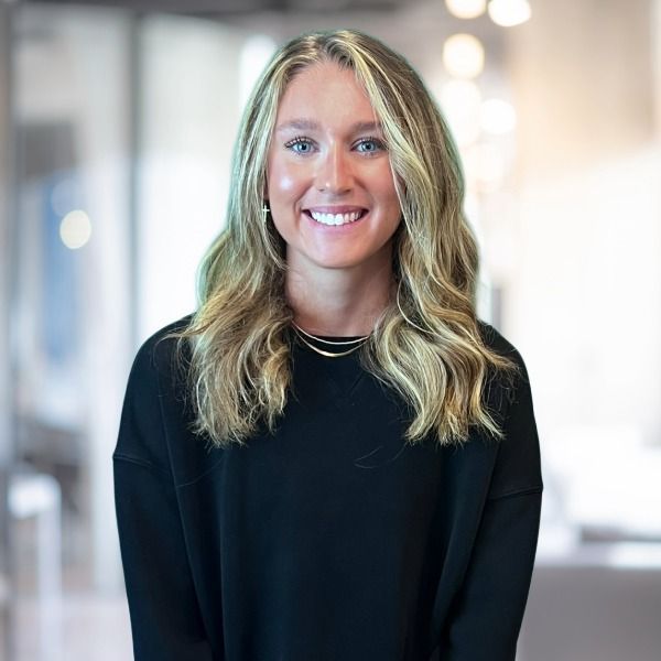 Woman with blonde hair smiling in a bright office setting, wearing a black sweater.
