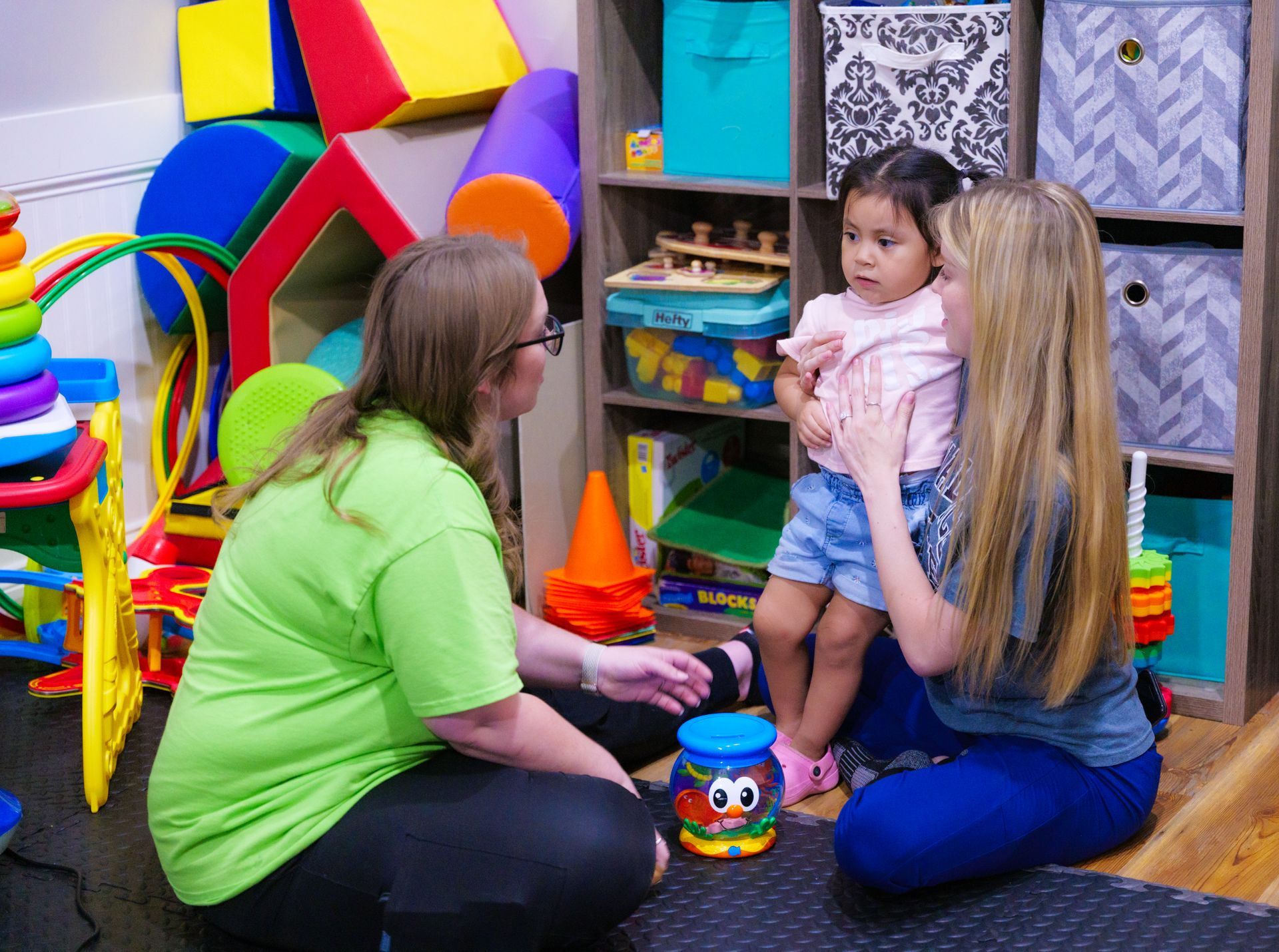 Two women are sitting on the floor talking to a baby in a play room.