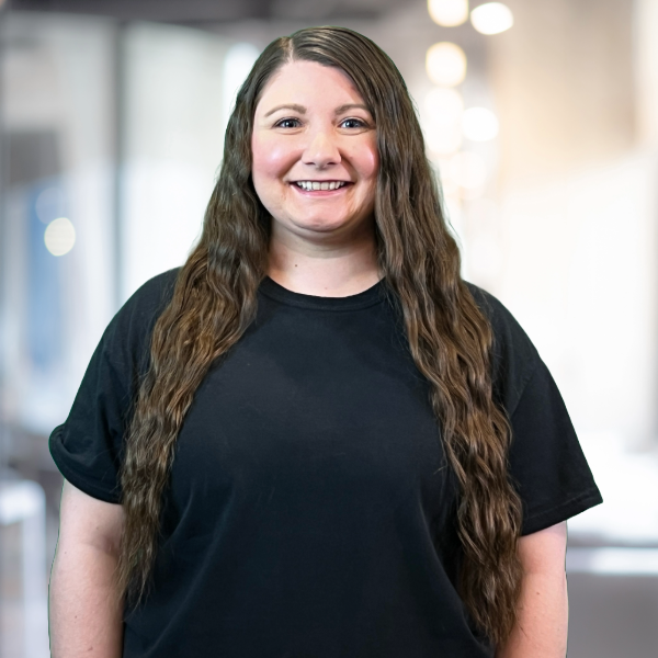 Woman with long brown hair smiles, wearing a black shirt, in a blurred office setting.