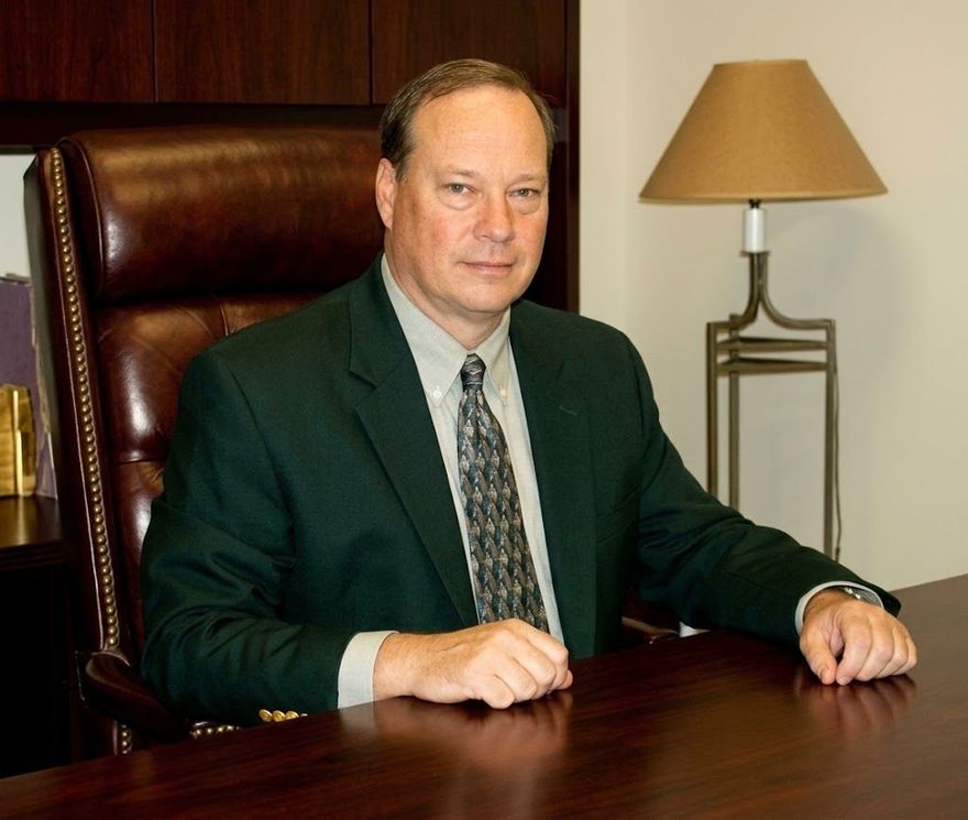 Man in a dark green suit seated at a wooden desk in an office, with a lamp and leather chair behind him.