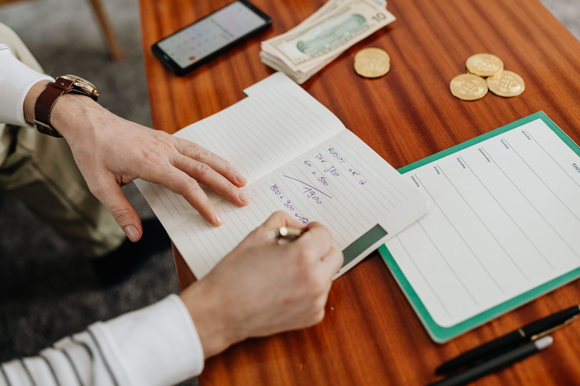 Hands writing in a notebook beside a tablet, coins, and a green-framed budget sheet on a wooden table