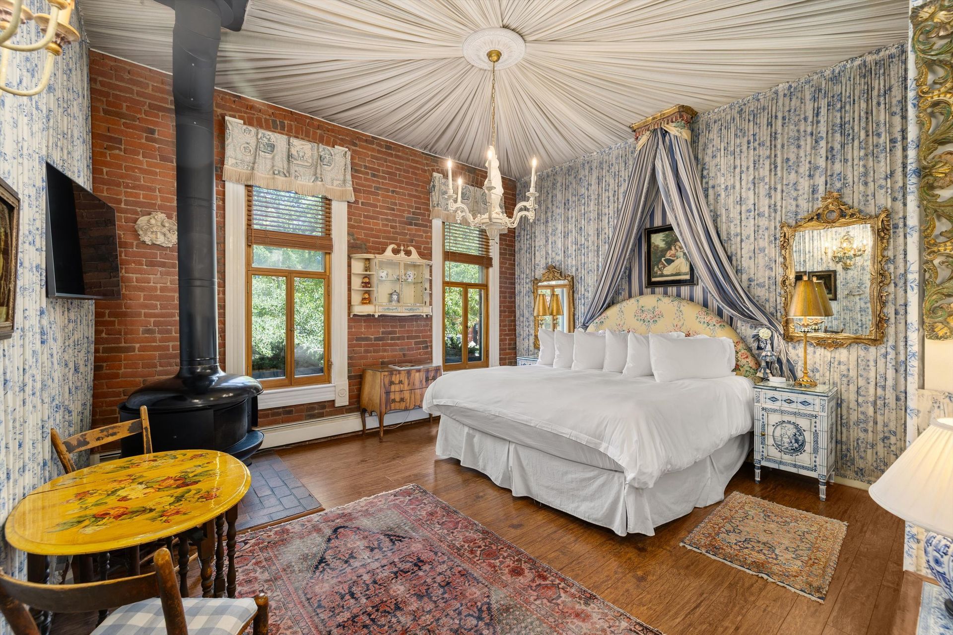 Bedroom with exposed brick wall, bed, chandelier, and a small wood-burning stove.