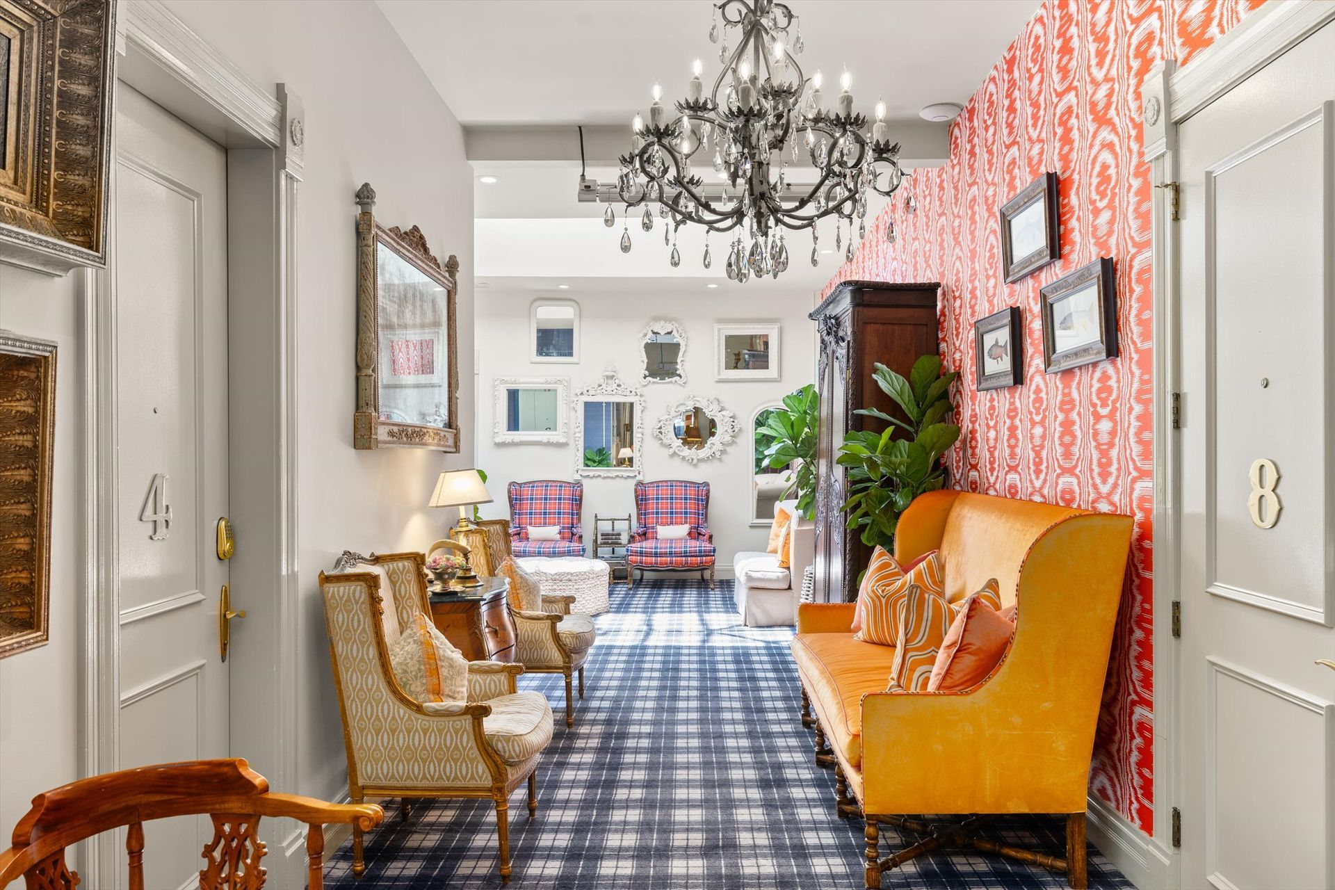 Hallway with ornate decor: chairs, patterned rug, orange sofa, red patterned wallpaper, mirrors, chandelier.