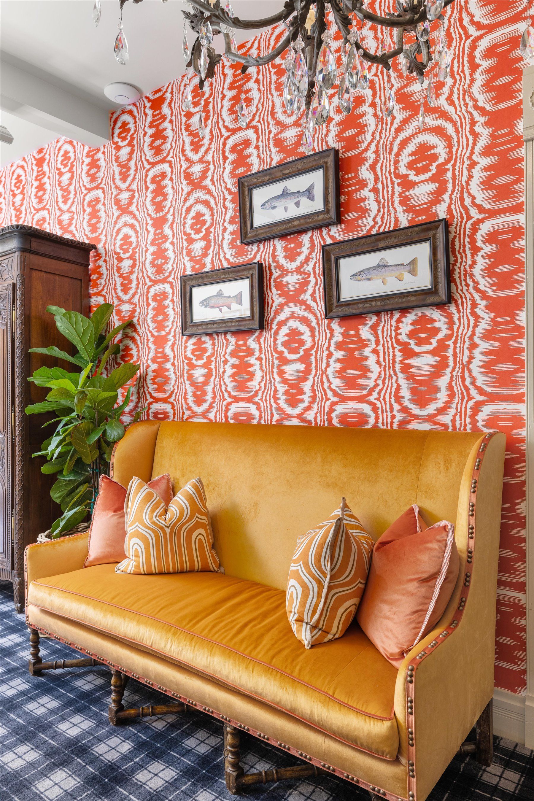 Orange velvet sofa with patterned pillows, against red and white wallpaper, three framed fish prints.